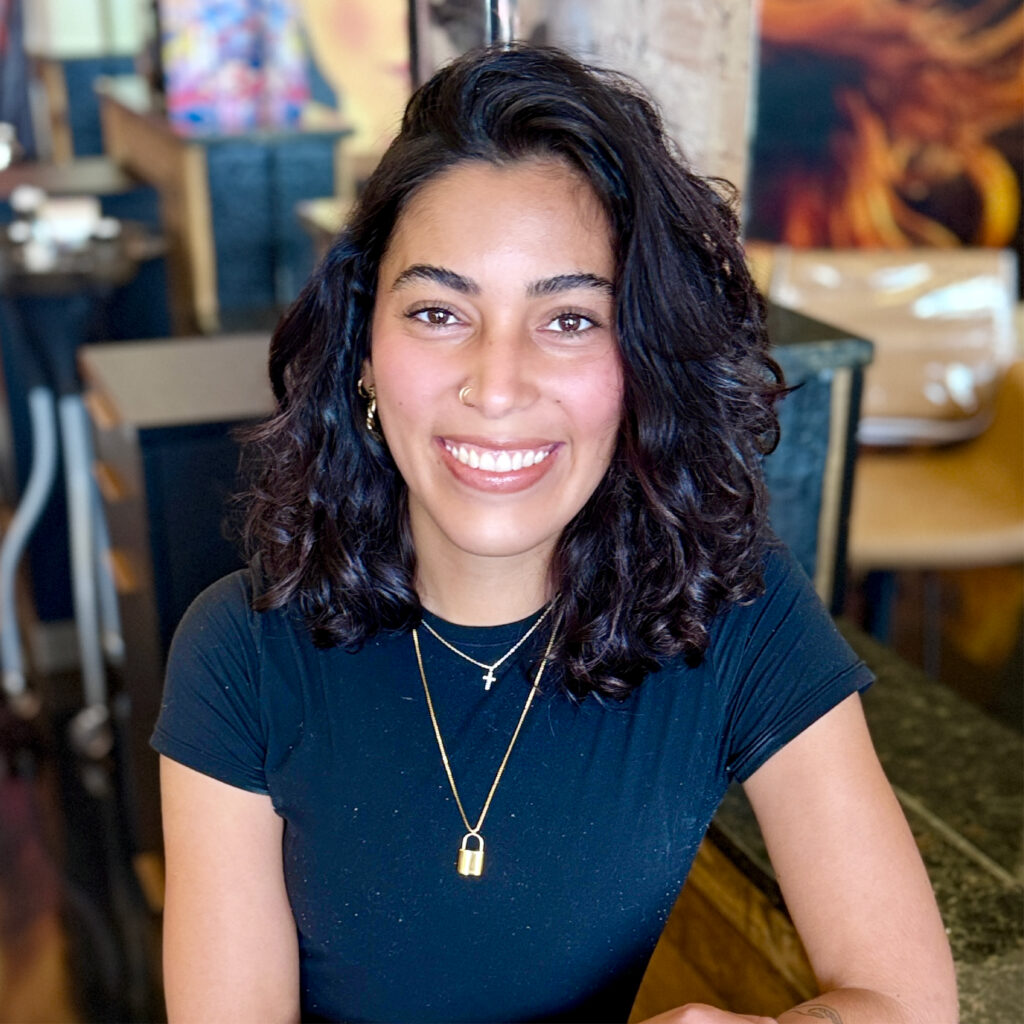 Amy Romulus, Tier 2 Hair Artist at J. Meraki Beauty Emporium, smiling and seated at a styling station.